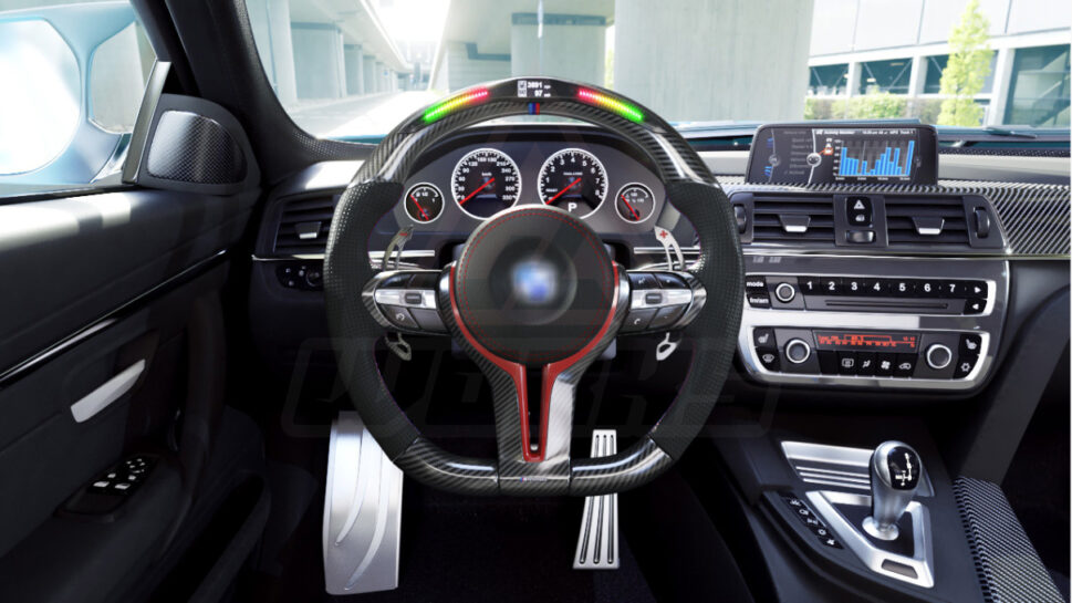 Front view of a BMW F chassis, M sport steering wheel in carbon fiber, hollow design paddle shifters, a mix of red and M color accents, black perforated leather and led shift lights